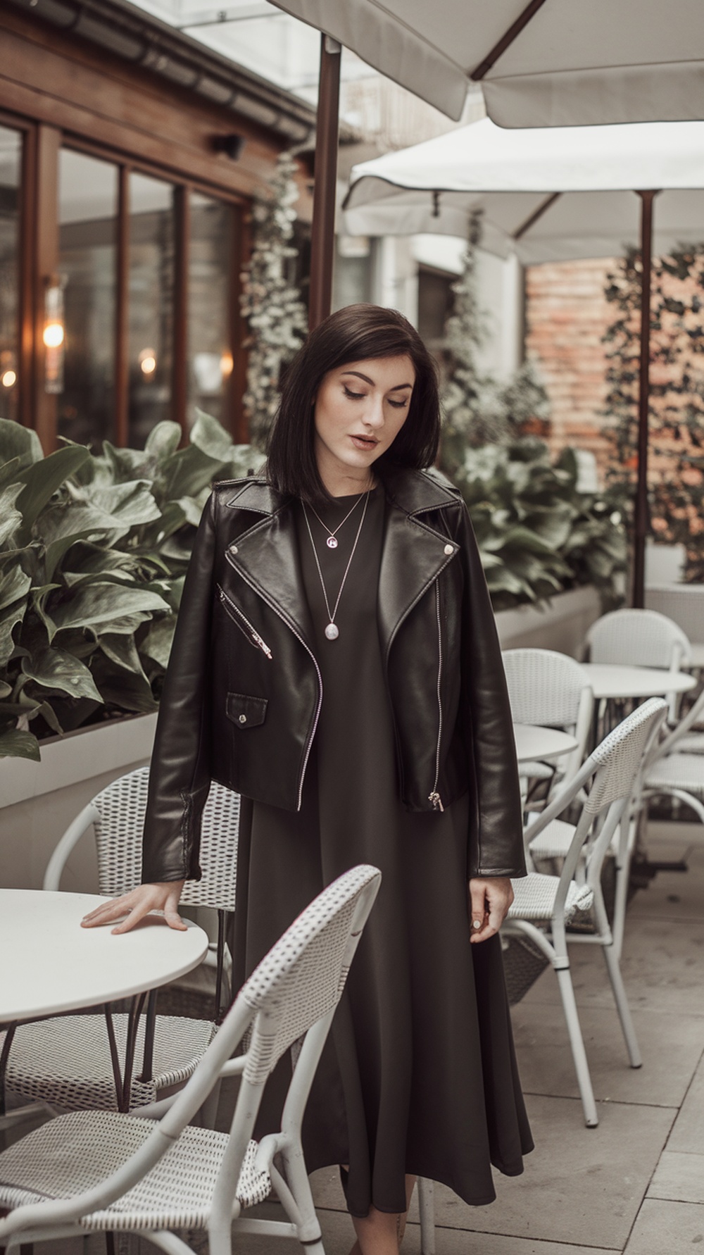 A woman wearing a black leather jacket over a midi dress, standing in a stylish outdoor café setting.